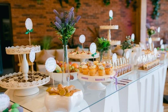 A beautifully arranged modern wedding dessert counter featuring frosted cupcakes, pastries, chocolate tarts, and fruit platters on elegant white tiered stands. Decorated with lavender floral arrangements, this premium dessert catering setup in Kolkata is provided by Food Chakra.