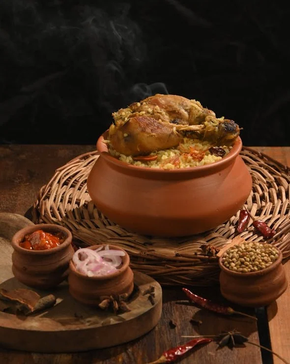 Steaming hot chicken biryani topped with a large roasted leg piece, served in a traditional clay handi on a woven mat. Accompanied by side bowls of raita, sliced onions, and whole spices, showcasing premium wedding catering services in Kolkata by Food Chakra.
