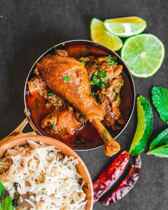A rich, authentic Indian chicken curry with a prominent drumstick served in a brass bowl, accompanied by aromatic basmati rice, mint leaves, and lime slices from Food Chakra catering in Kolkata. | Image for Food Chakra – Trusted Catering Services in Kolkata
