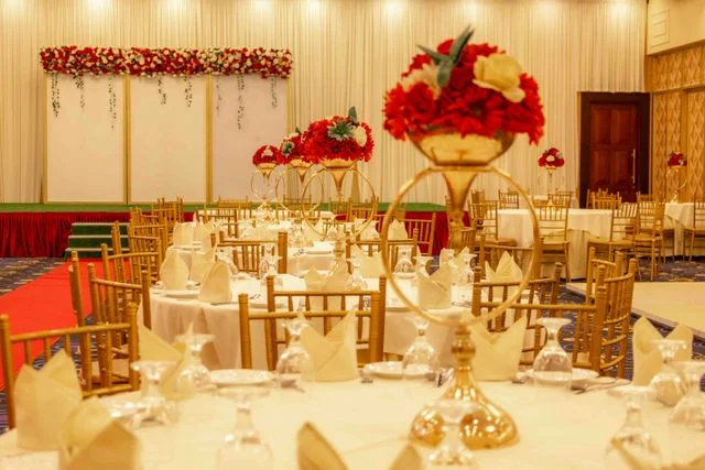 A luxurious wedding reception hall banquet setup featuring round dining tables with white linens, gold Chiavari chairs, and tall gold floral centerpieces with red roses. The grand venue arrangement showcases premium wedding catering and hospitality services in Kolkata by Food Chakra.