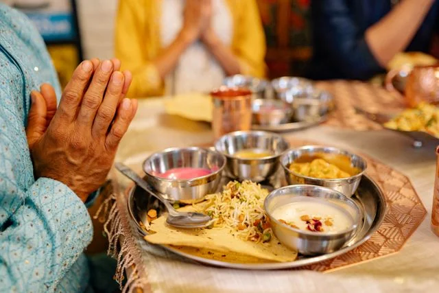 Close-up of a guest in traditional attire with hands folded in a prayer gesture before partaking in a festive feast. A meticulously arranged thali sits on a patterned placemat, containing steel katoris (bowls) filled with authentic curries, a vibrant pink chutney, creamy payesh/kheer topped with rose petals, seasoned rice, and crisp papad. This image perfectly captures Food Chakra's dedication to preserving traditional customs and authentic food culture for weddings and sacred rituals in Kolkata. | Food Chakra Catering Services for Religious Function in Kolkata