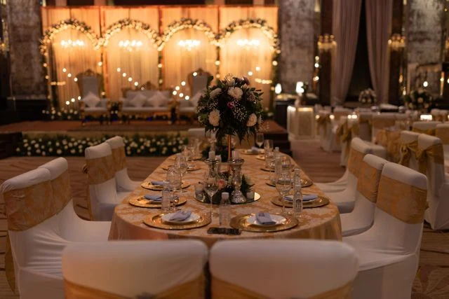 A luxurious oval VIP dining table set with gold charger plates, fine glassware, and floral centerpieces inside a grand wedding reception venue. The background features a brilliantly lit wedding stage with floral arches, representing premium event hospitality and catering in Kolkata by Food Chakra.